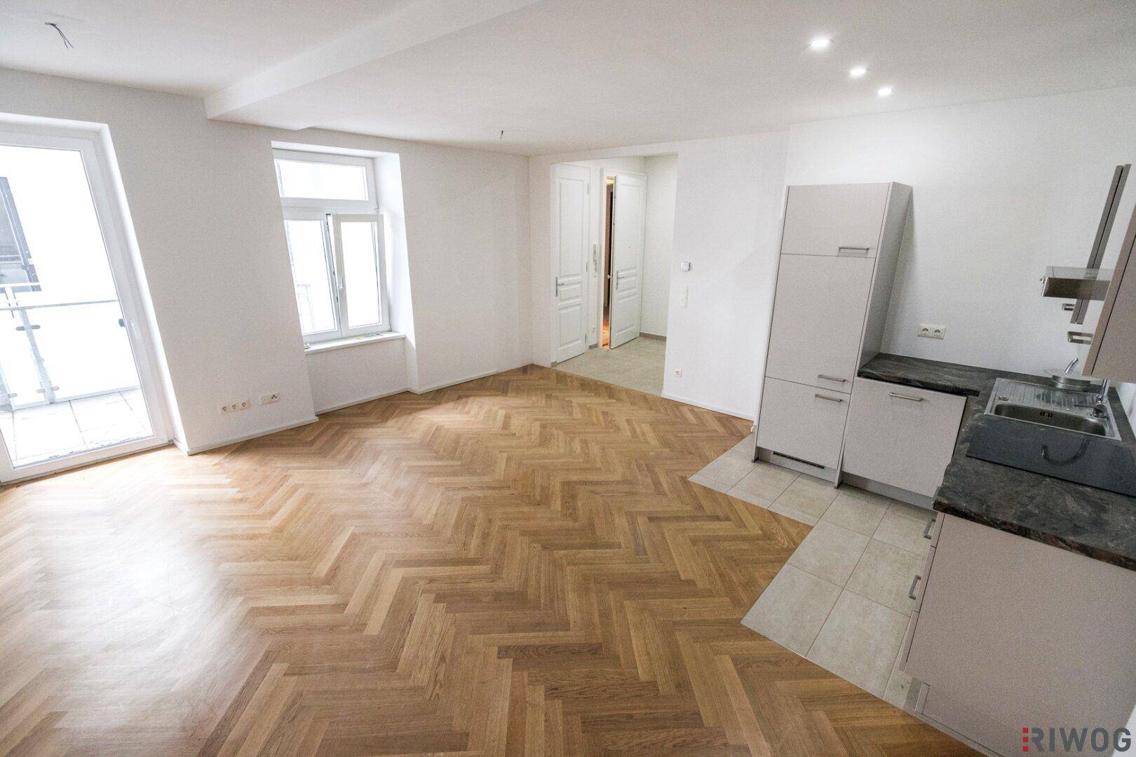 Heller Wohn-Essbereich mit Fischgrätparkett, Zugang zum Balkon und integrierter Küchenzeile.
