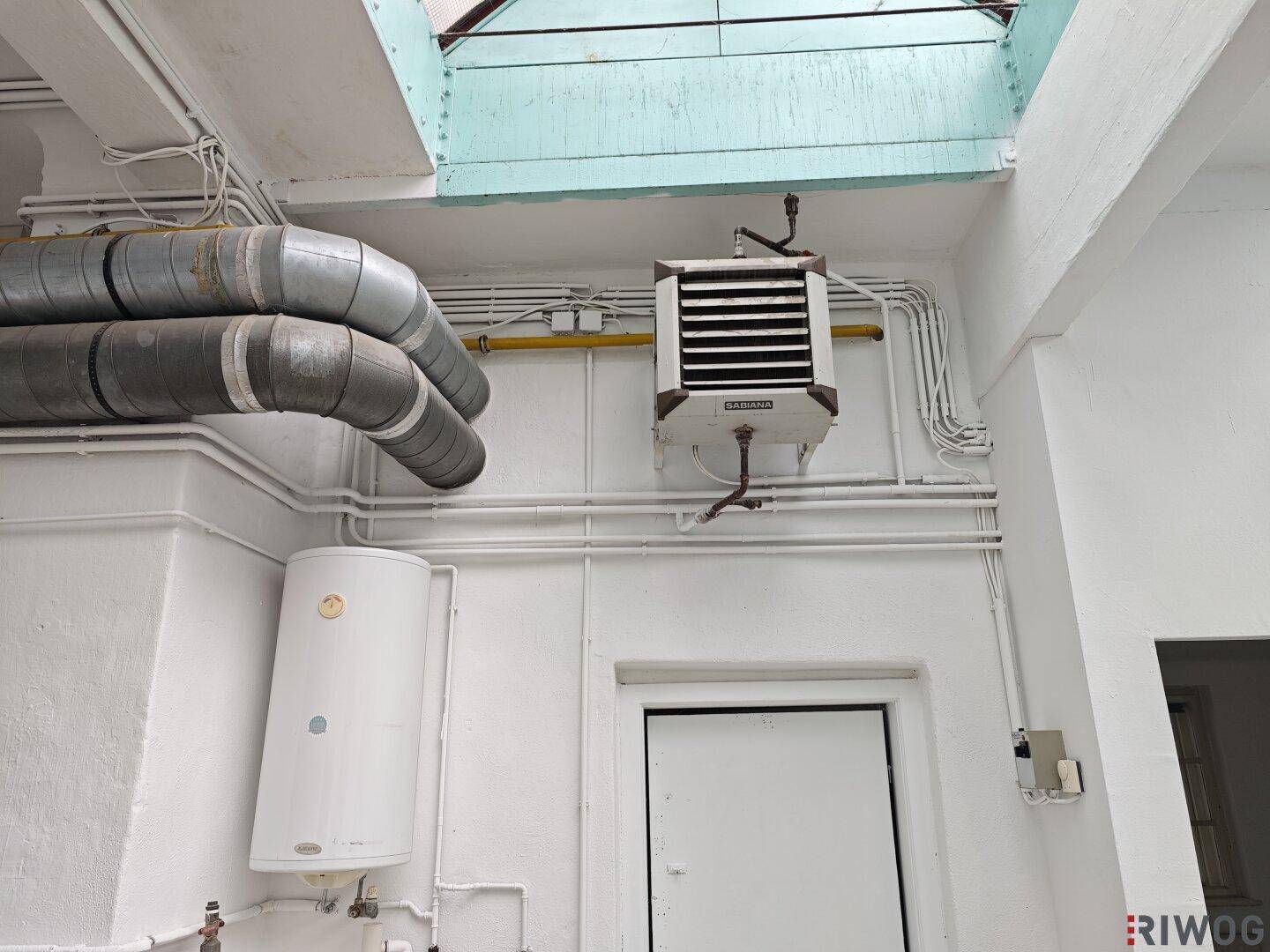 Detailansicht von Heizungsrohren, Warmwasserspeicher und einer Wandheizung unter dem Glasdach.