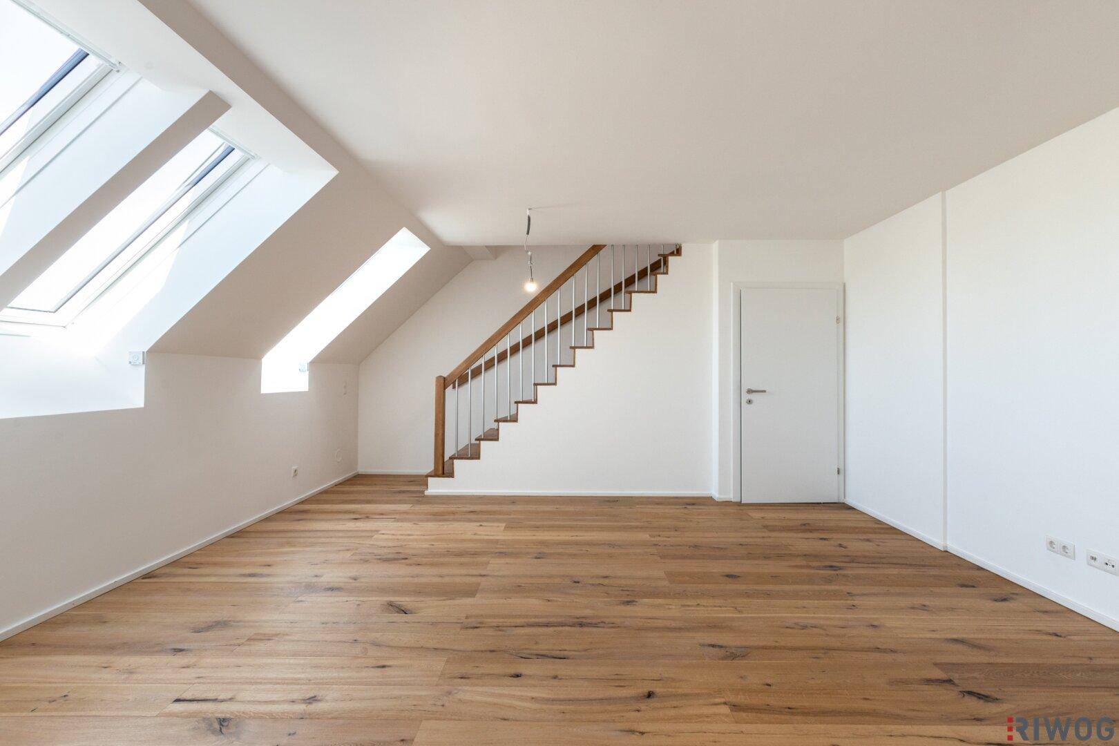 Heller Raum mit Holzboden, Treppe und Dachfenstern, der zu weiteren Ebenen führt.