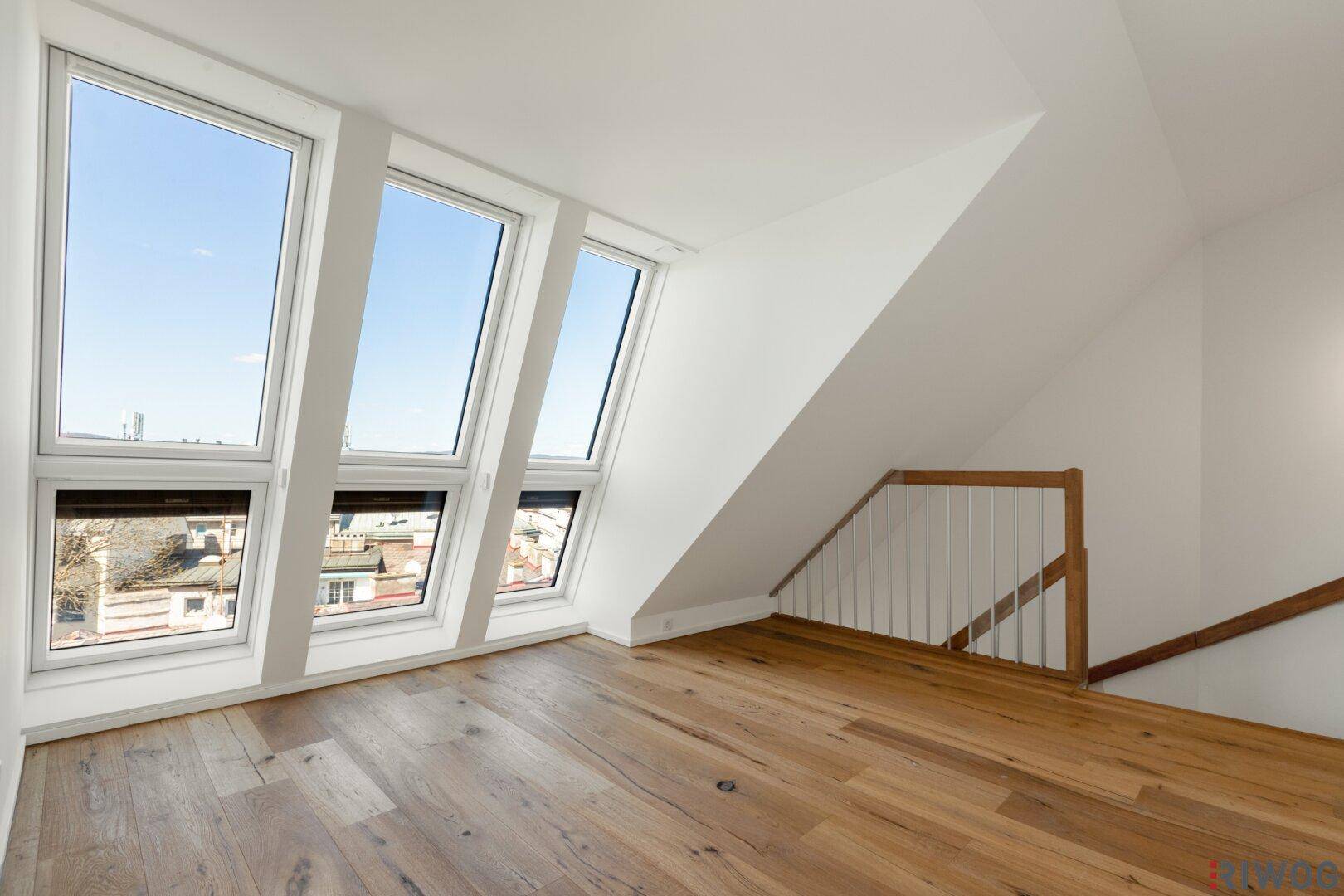 Heller leerer Raum mit Holzboden, großen Fenstern und Zugang zur Treppe.