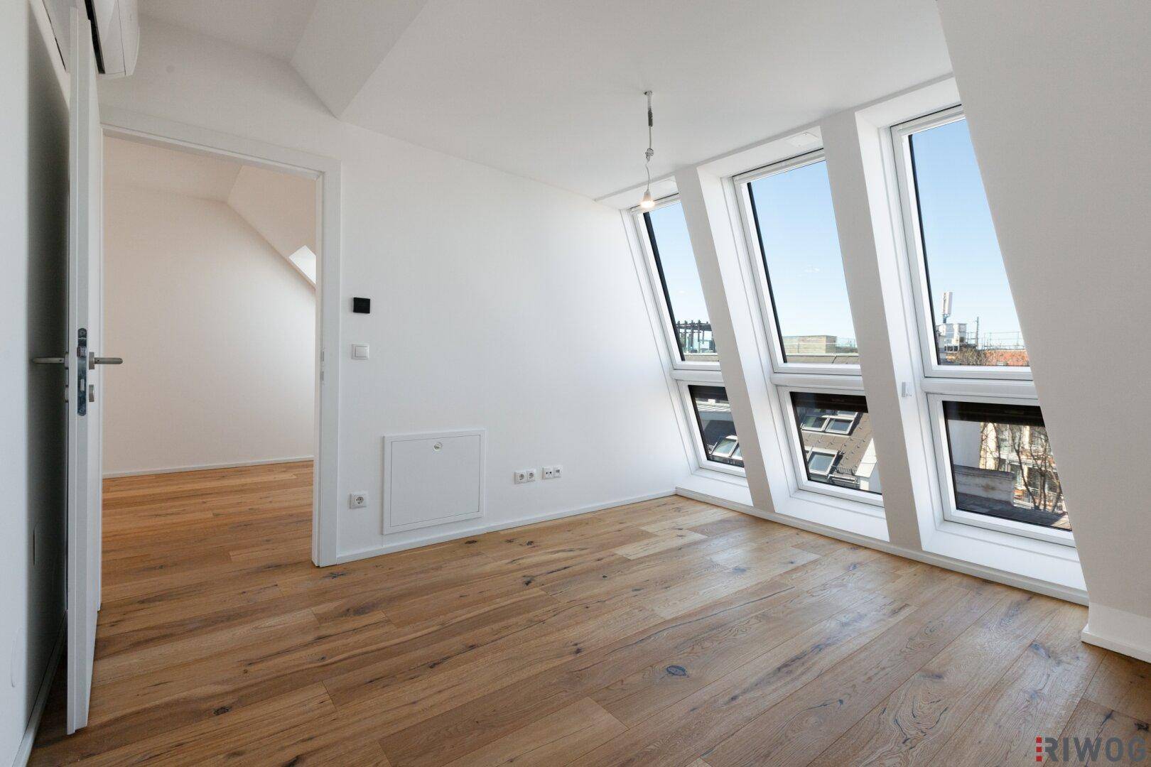 Leerer Raum mit Holzboden und großen Fenstern, die viel Tageslicht hereinlassen.
