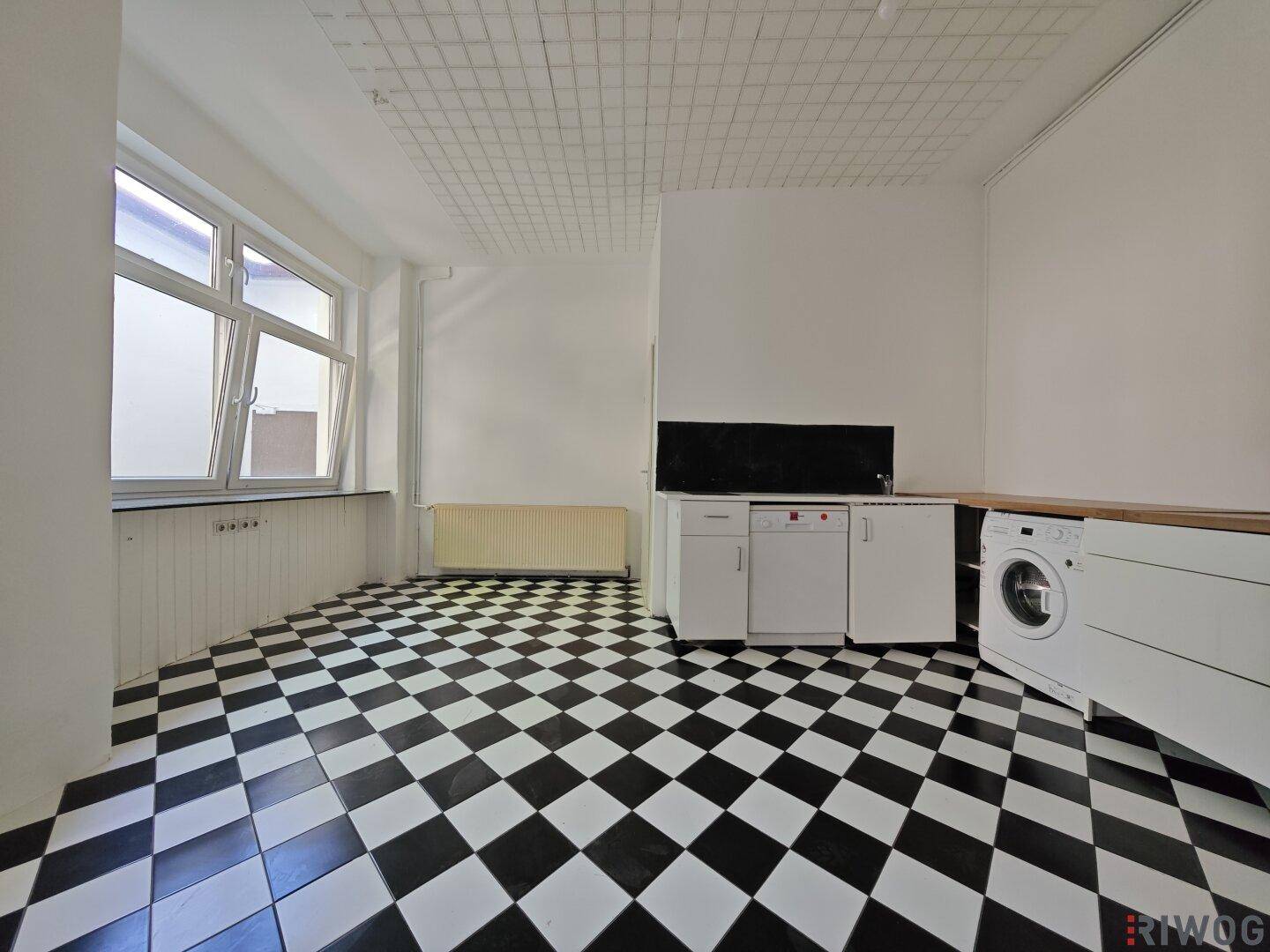 Raum mit schwarz-weiß kariertem Fliesenboden, Fenster und einer Küchenzeile mit Waschmaschine.