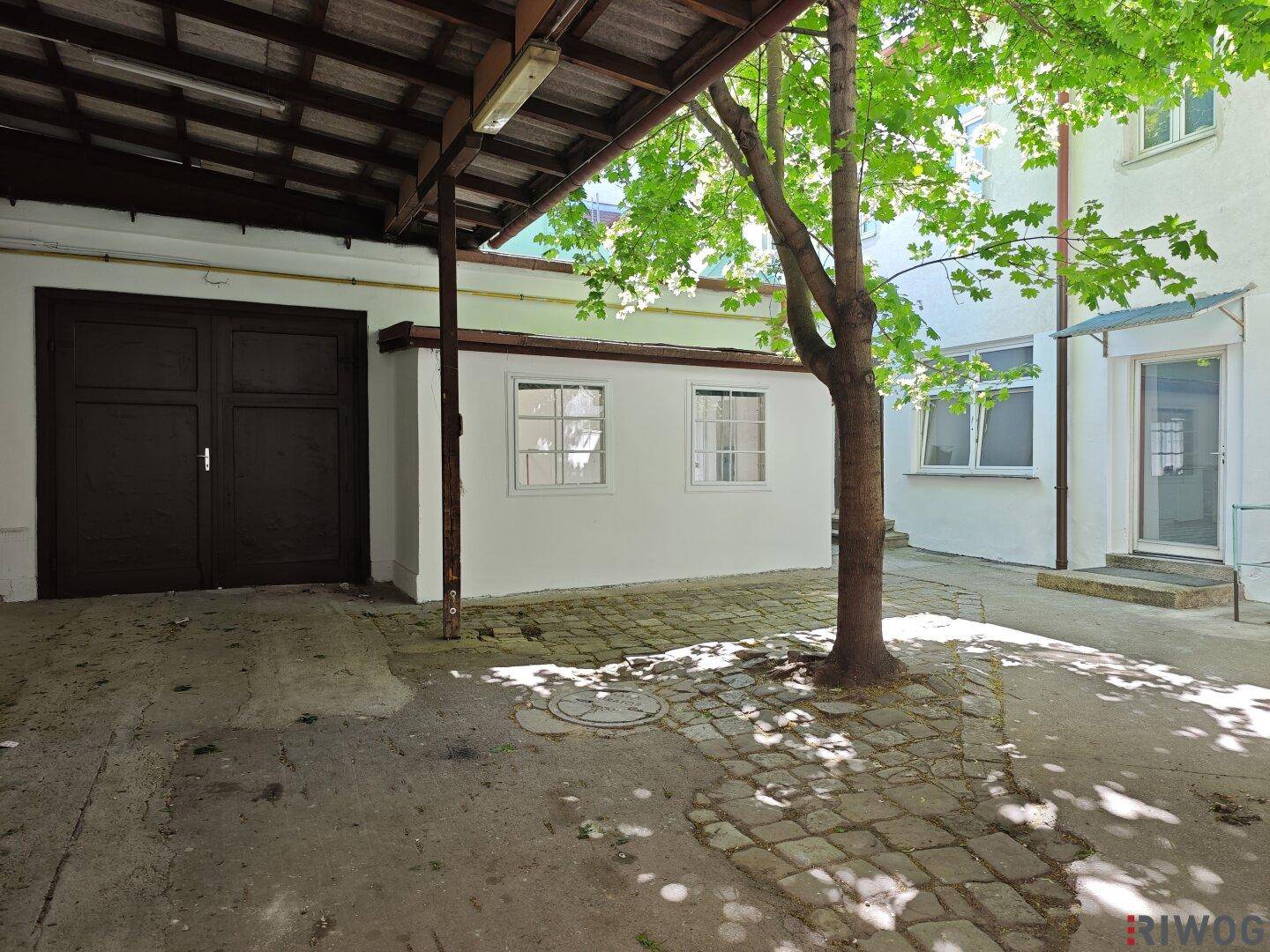 Innenhof mit Baum, überdachtem Stellplatz und einer Garageneinfahrt, umgeben von Gebäuden.