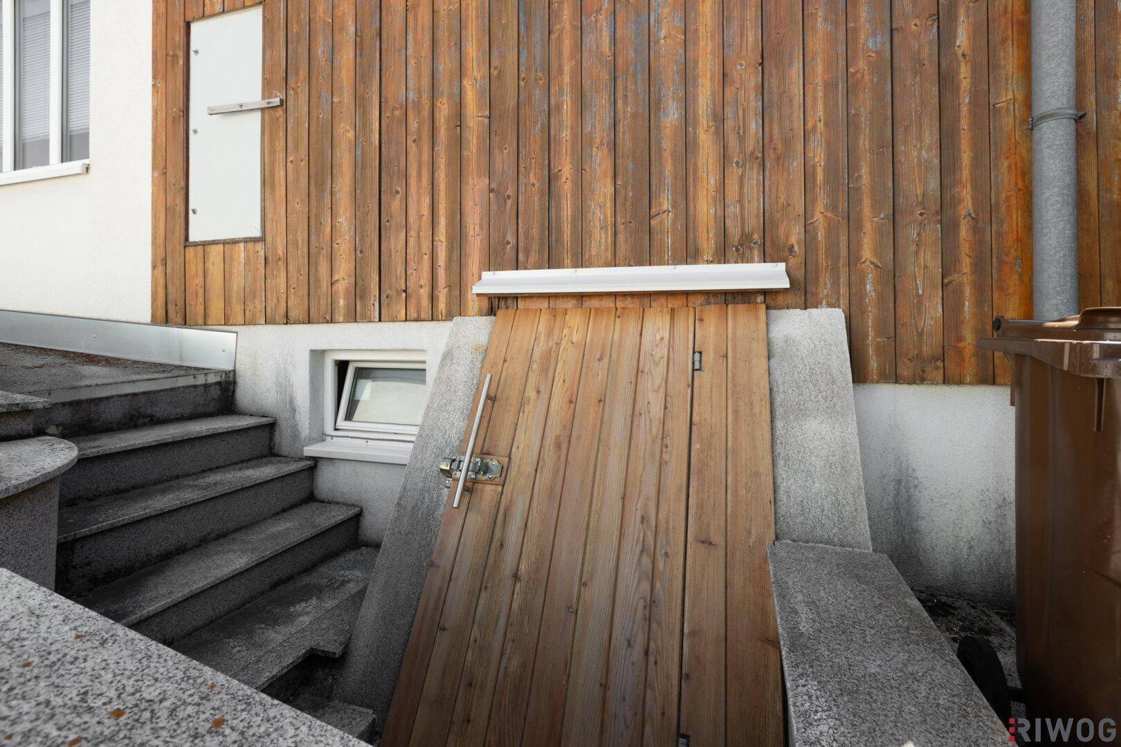 Außenansicht des Kellerzugangs mit Holzklappe und Steintreppe, die zum Untergeschoss führt.