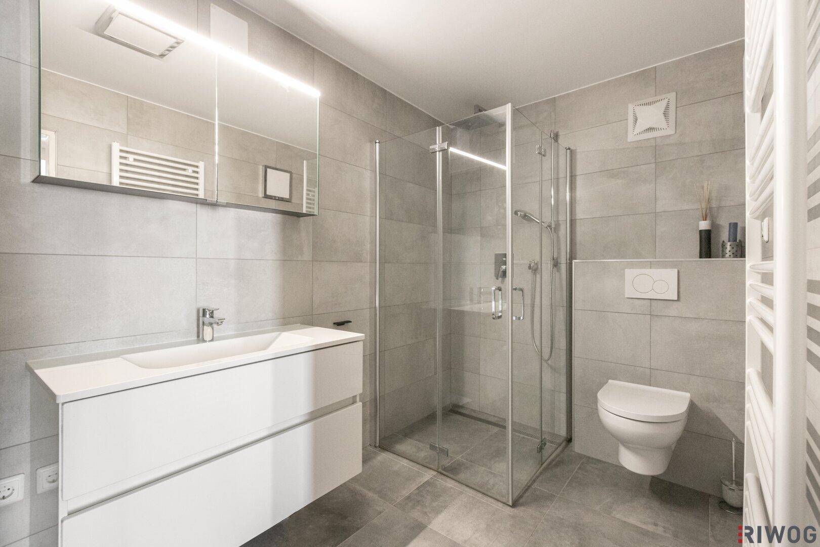 Modernes Badezimmer mit großen grauen Fliesen, Duschkabine und wandhängendem WC.