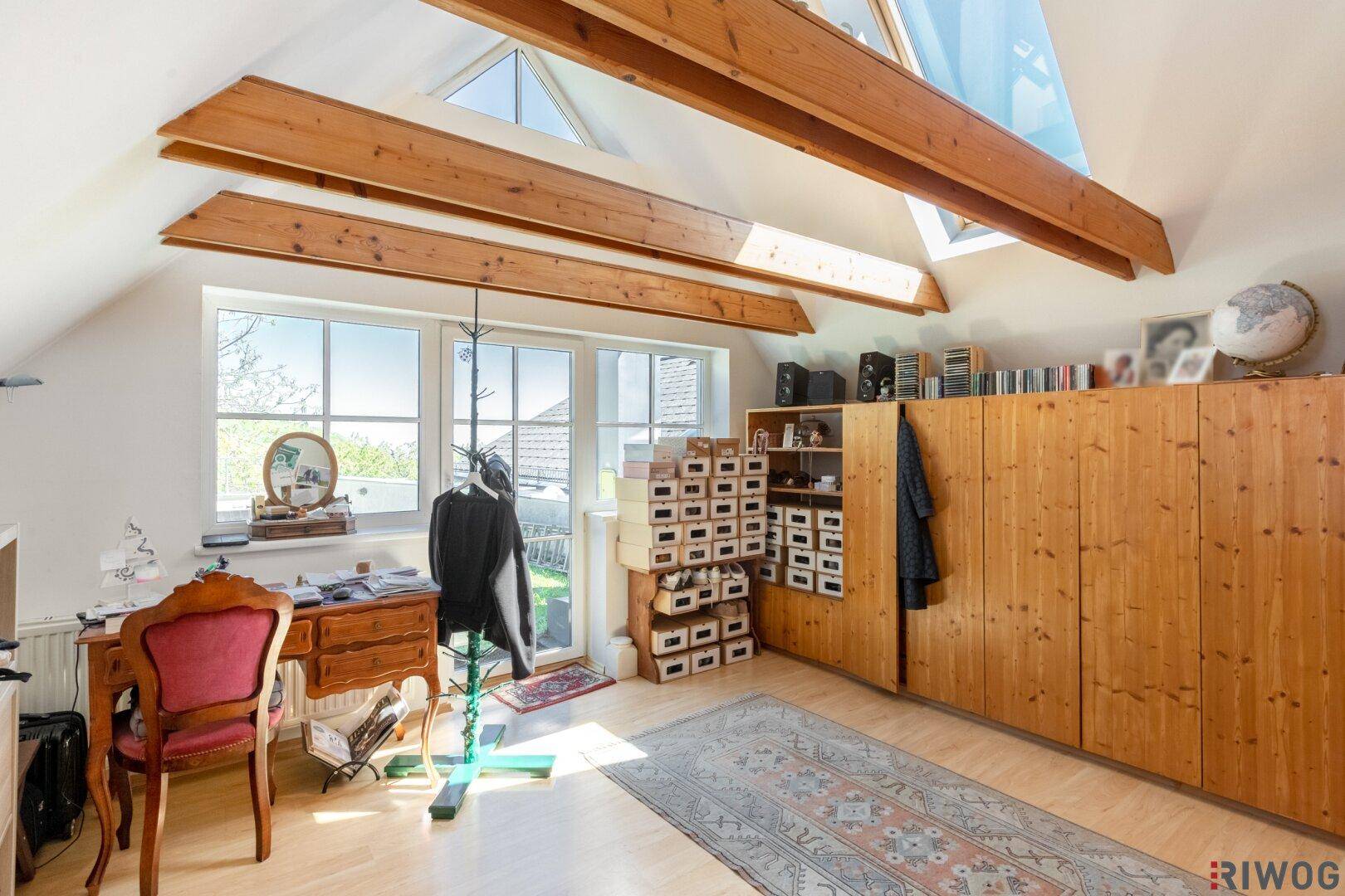 Helles Arbeitszimmer im Dachgeschoss mit Holzbalken, großem Fenster und Zugang zum Außenbereich.