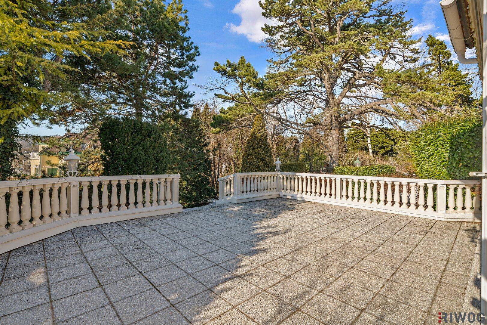 Große gepflasterte Terrasse mit Balustrade, umgeben von hohen Bäumen und Grünflächen.