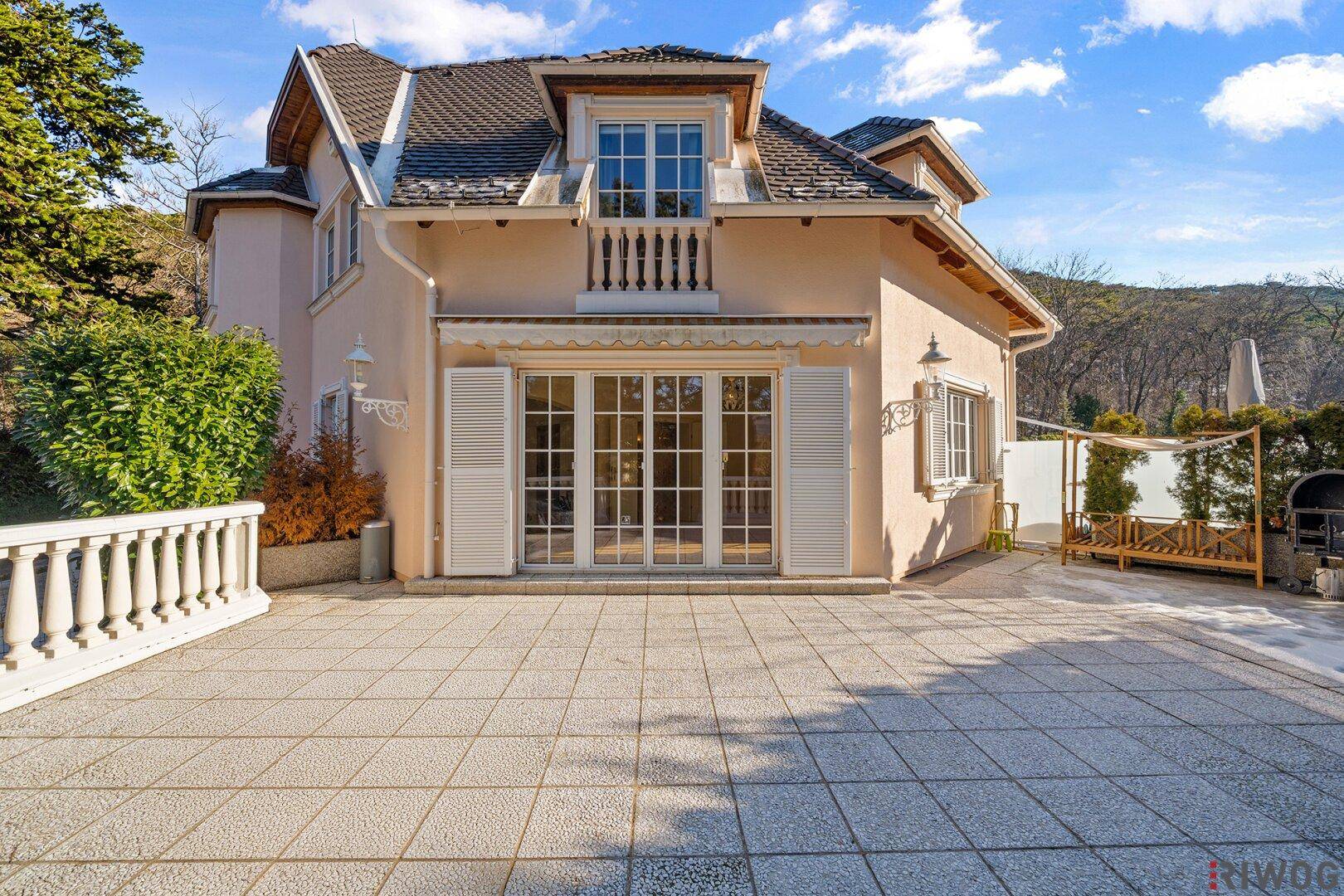 Großzügige gepflasterte Terrasse vor dem Haus mit Zugang zu den Innenräumen und einem Balkon darüber.