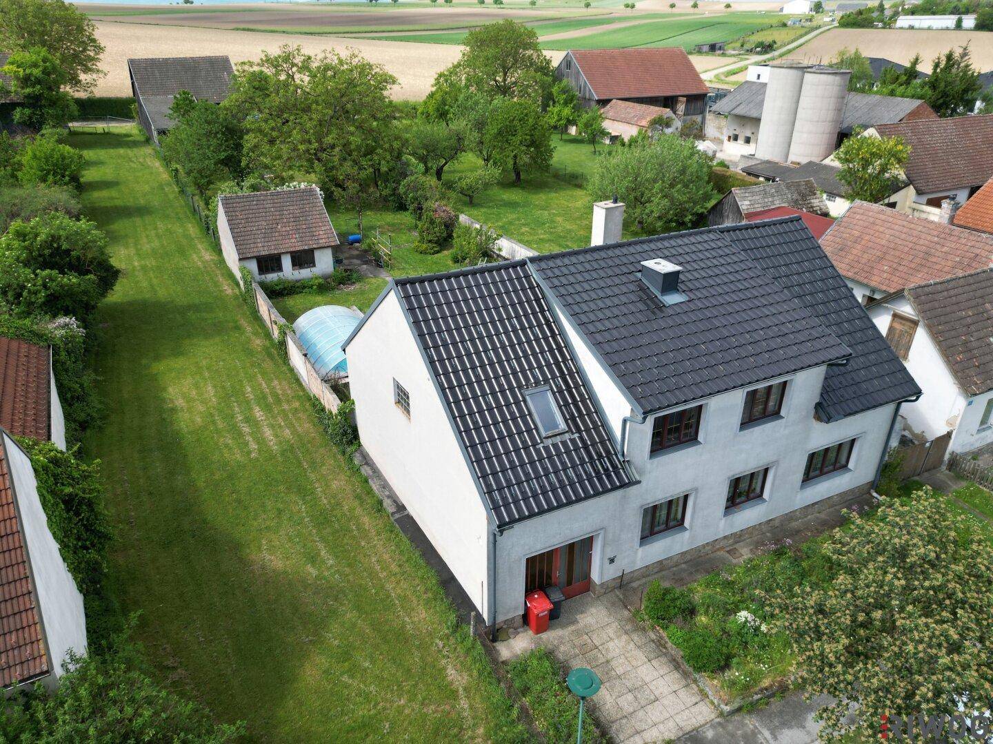 Vorderansicht eines Einfamilienhauses mit dunklem Ziegeldach und kleinem Vorgarten, umgeben von Grünflächen.