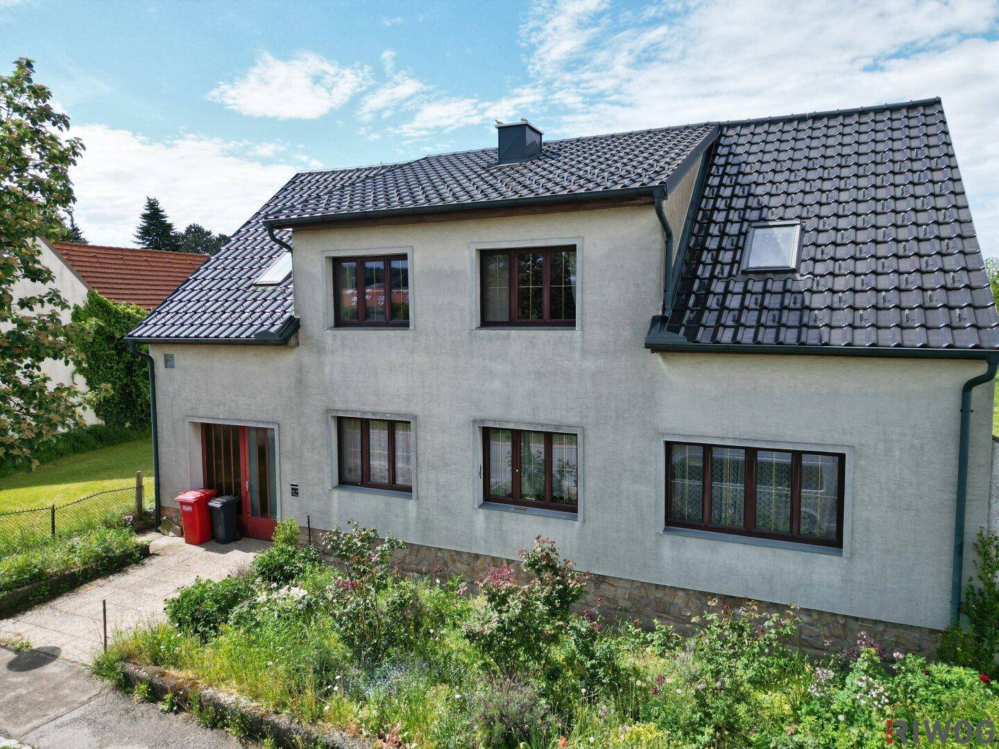 Fassade eines Wohnhauses mit weißem Putz, dunklem Ziegeldach und kleinen Fenstern im Vorgartenbereich.