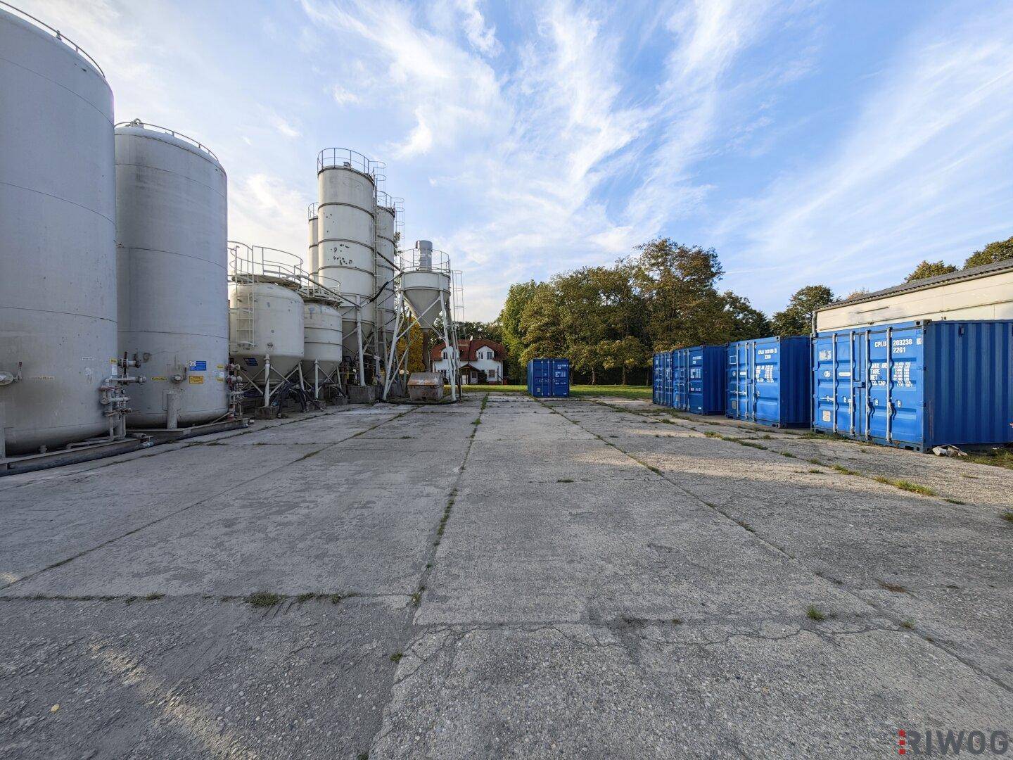 Nahaufnahme der großen Silos und blauen Lagercontainer auf dem weitläufigen, befestigten Hof des Geländes.