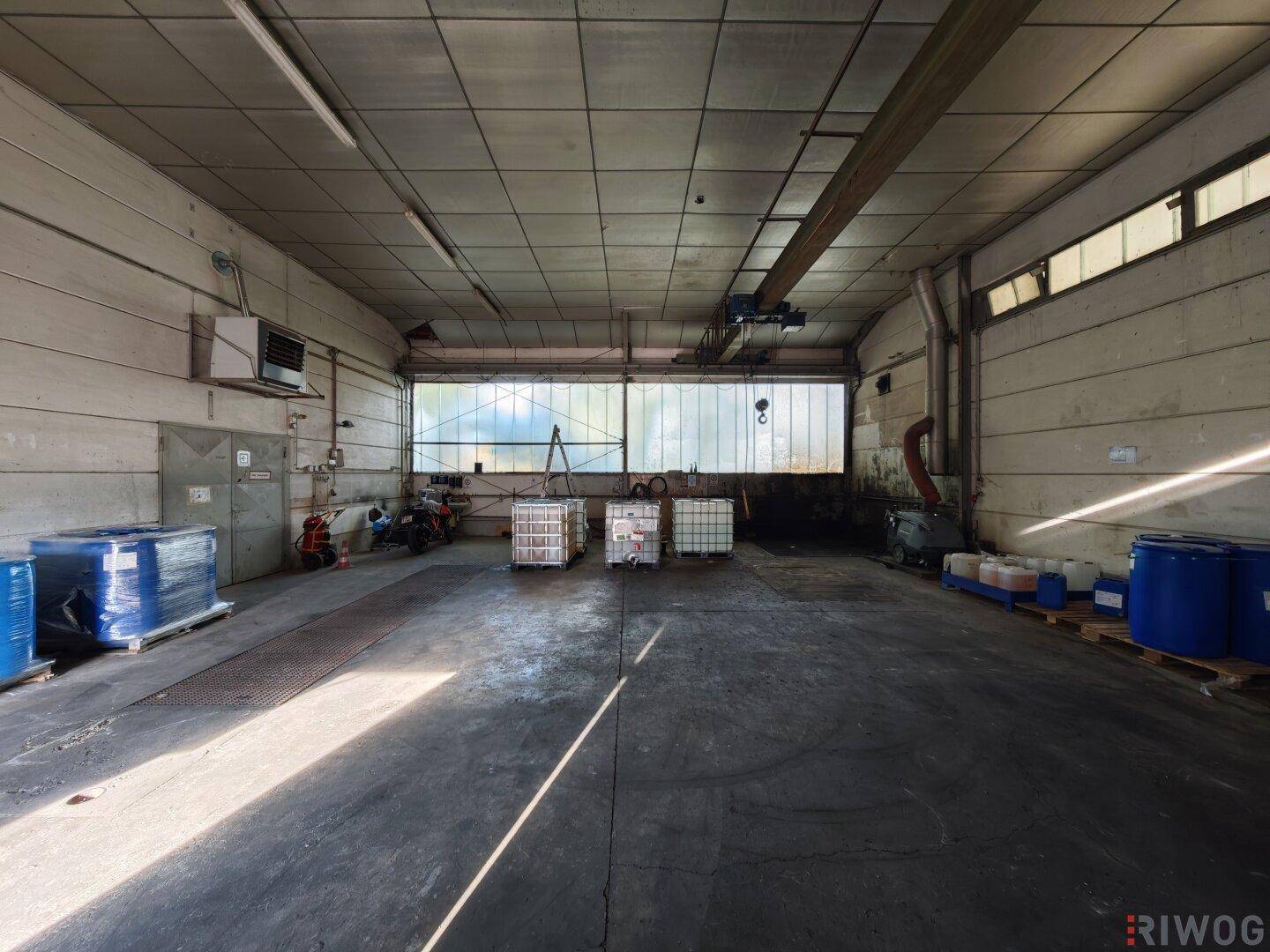 Geräumige Lagerhalle mit Betonboden, hoher Decke und Tageslicht durch ein großes Fenster im Hintergrund.