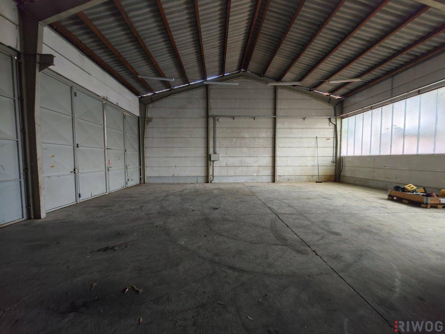 Leere Lagerhalle mit Betonboden, Wellblechdach und großen Rolltoren an der Seite für einfachen Zugang.