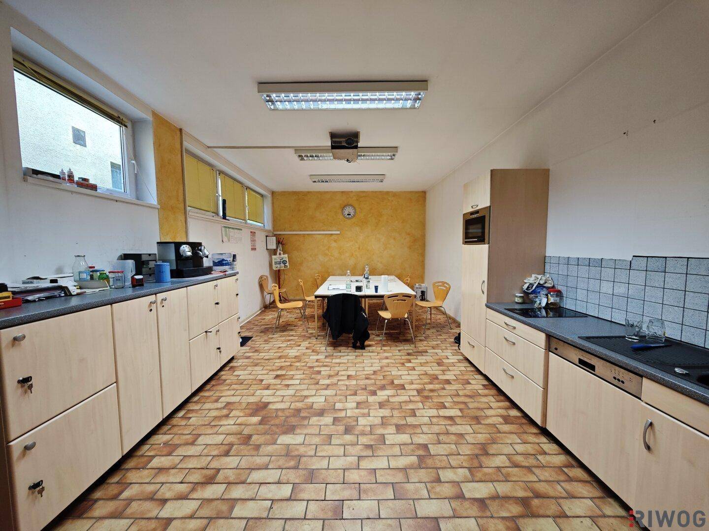 Mitarbeiterküche mit Terrakotta-Fliesenboden, Holzschränken, Esstisch und Fenstern für Tageslicht.