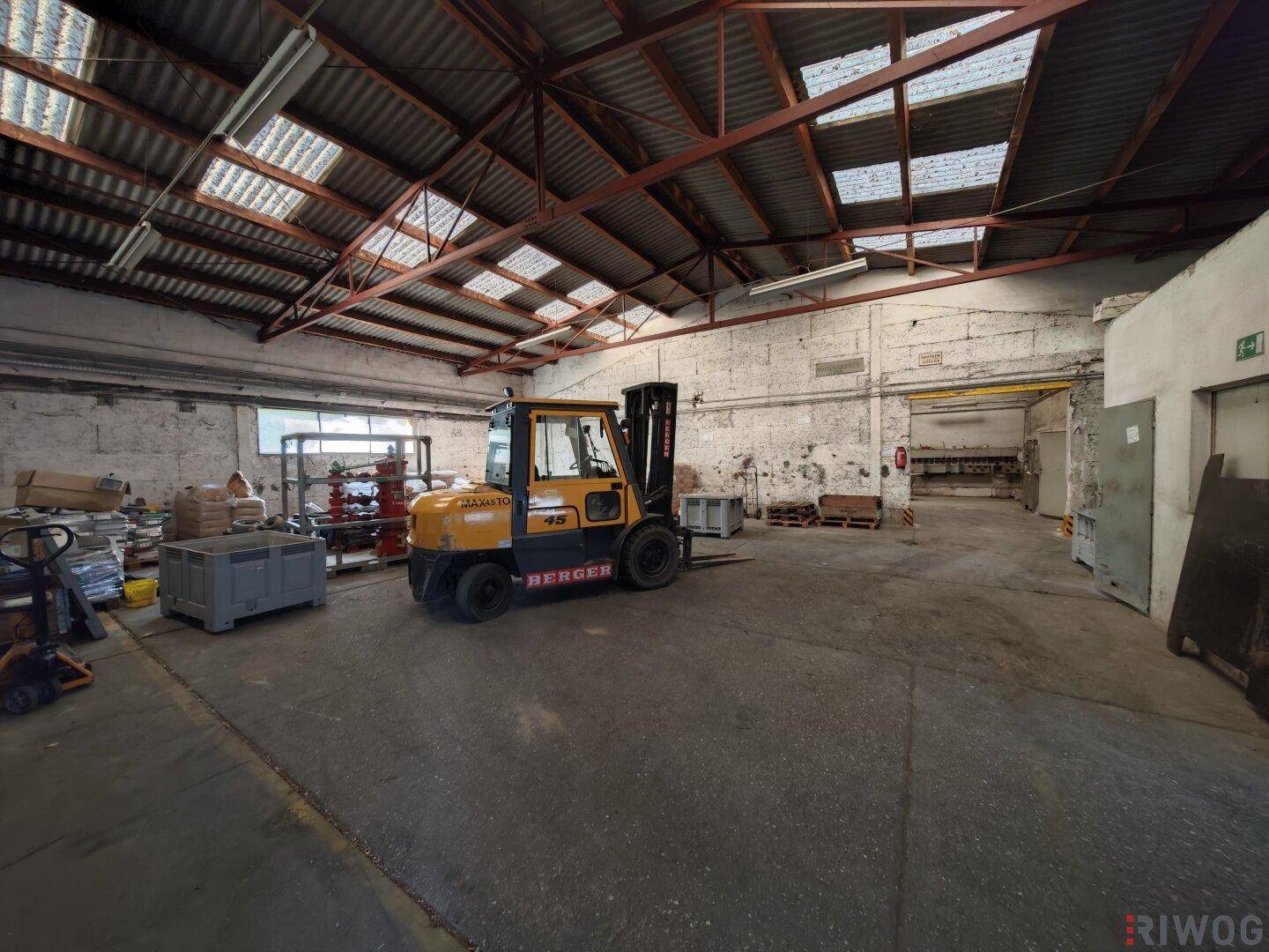 Große Lagerhalle mit Gabelstapler, Betonboden, hoher Decke und Oberlichtern für natürliche Beleuchtung.