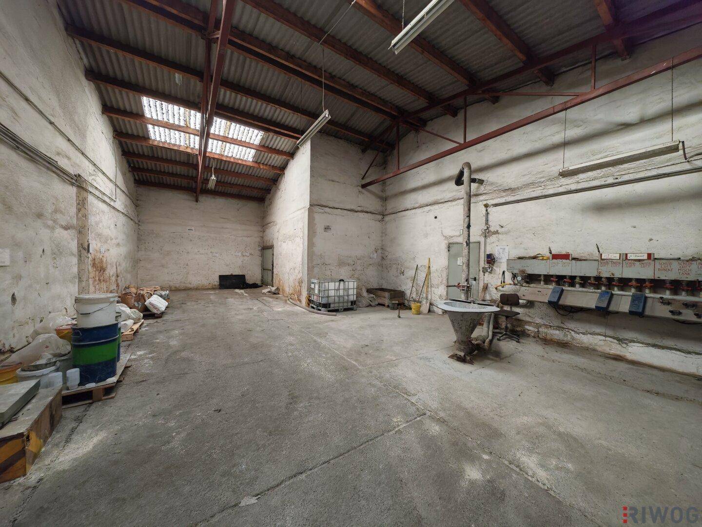 Geräumige Lagerhalle mit Betonboden, hoher Decke und Oberlichtern, bietet viel Platz für Materialien.