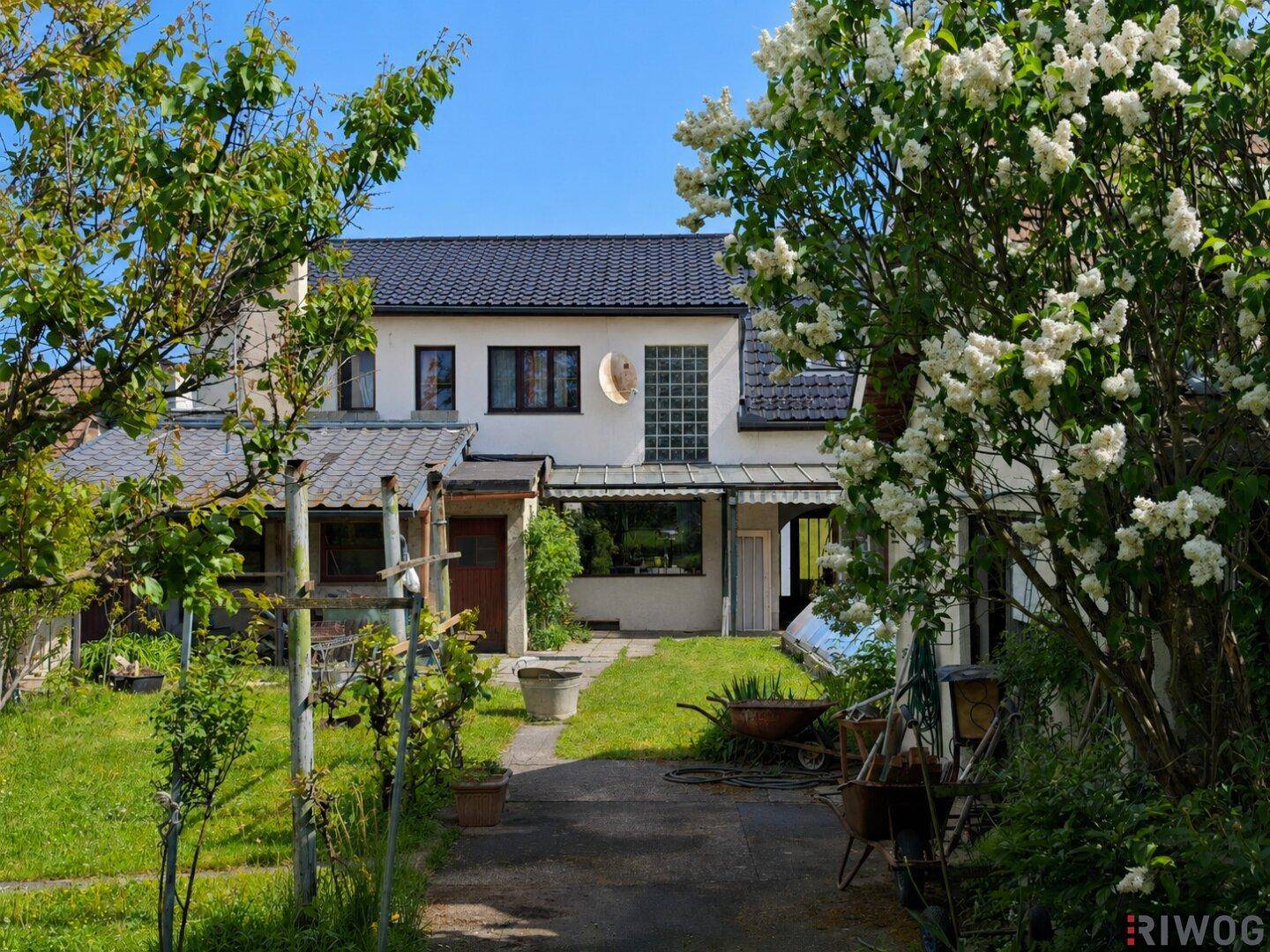 Hausfassade mit Gartenweg, umgeben von grünen Bäumen und blühenden Sträuchern unter blauem Himmel.