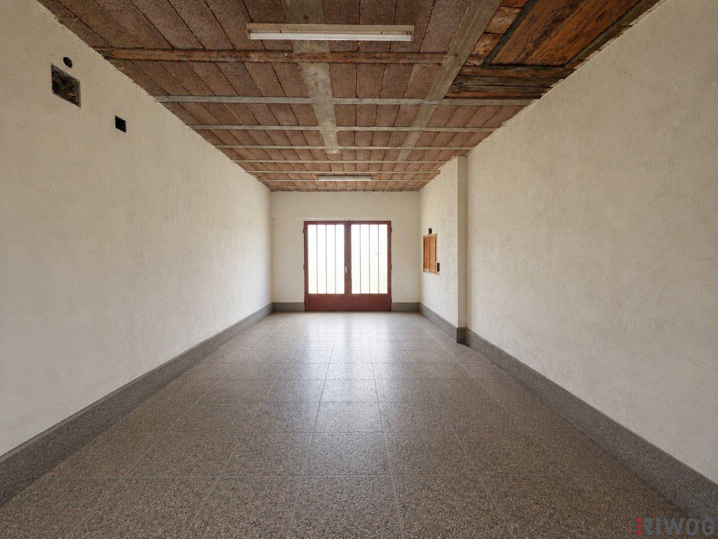 Leerer, langer Raum mit Holzbalkendecke und Terrazzoboden, bereit für eine neue Nutzung oder Renovierung.