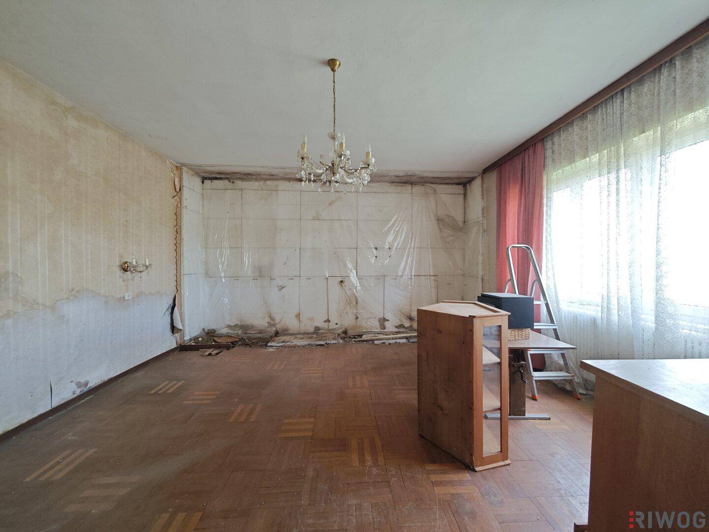 Renovierungsbedürftiger Raum mit Parkettboden und großem Fenster, bietet viel Tageslicht und Gestaltungspotenzial.