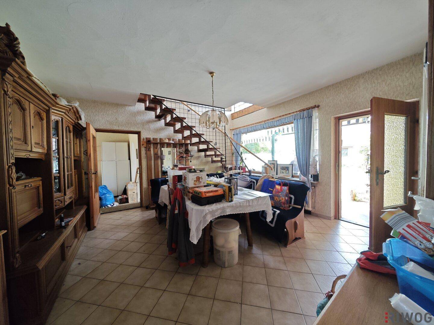 Geräumiger Wohn-Essbereich mit Fliesenboden, Holzmöbeln und offener Treppe, die in die obere Etage führt.