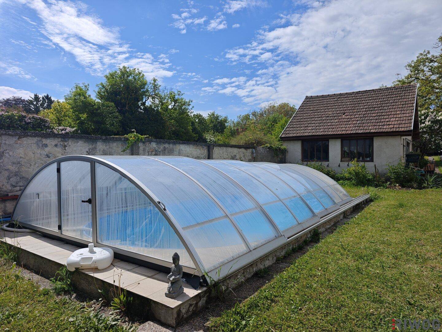 Großer Garten mit überdachtem Swimmingpool und einem kleinen Nebengebäude unter blauem Himmel.