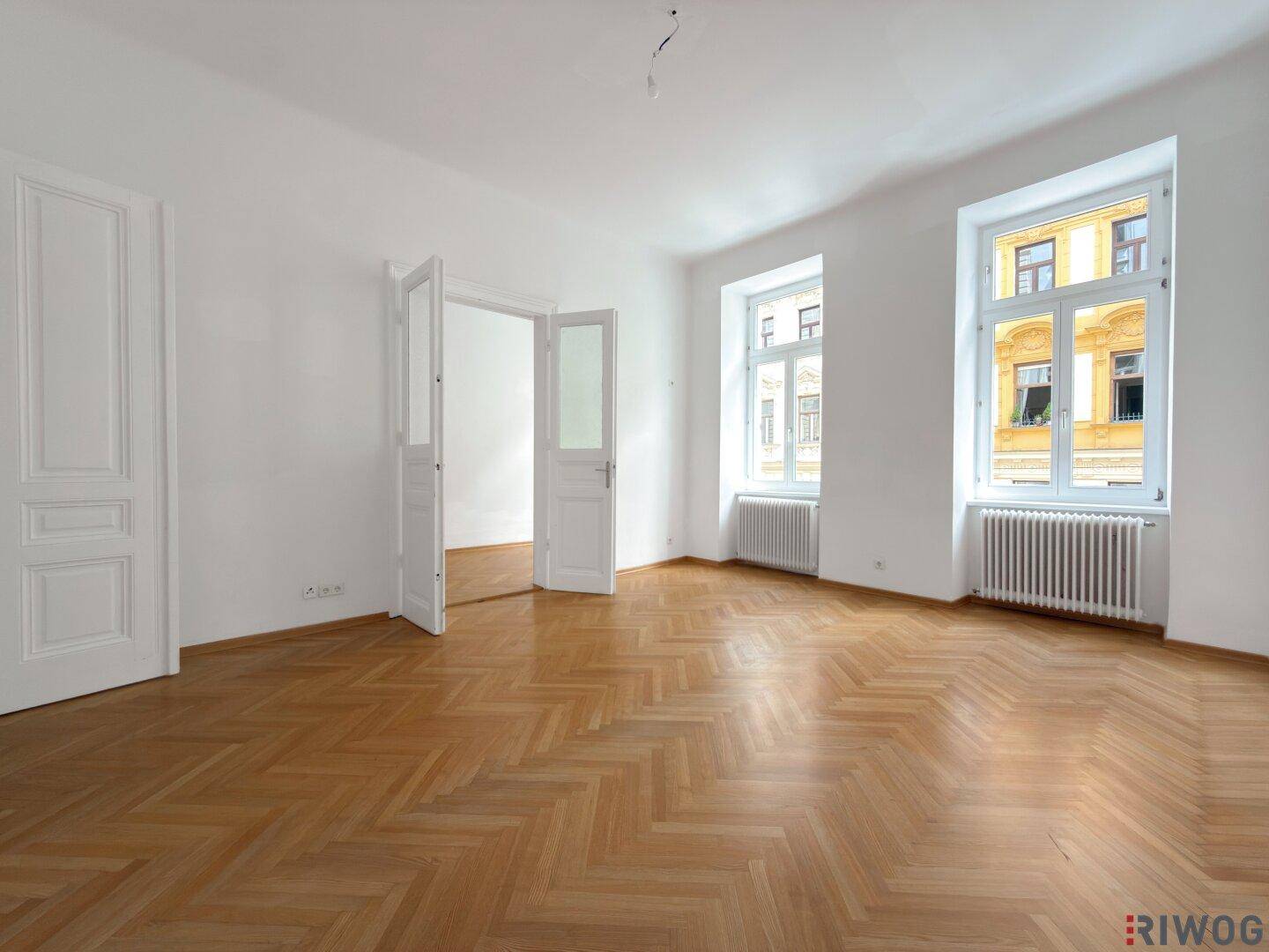 Leeres Zimmer mit hellem Fischgrätparkett, hohen Decken und zwei großen Fenstern.