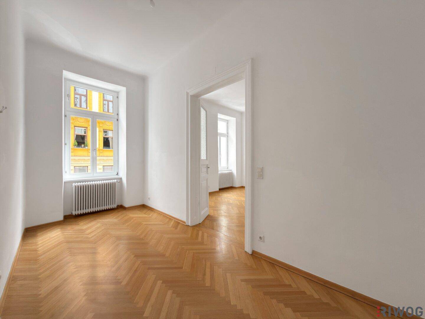 Helles Zimmer mit Fischgrätparkett und einem Fenster, das einen Blick nach draußen ermöglicht.