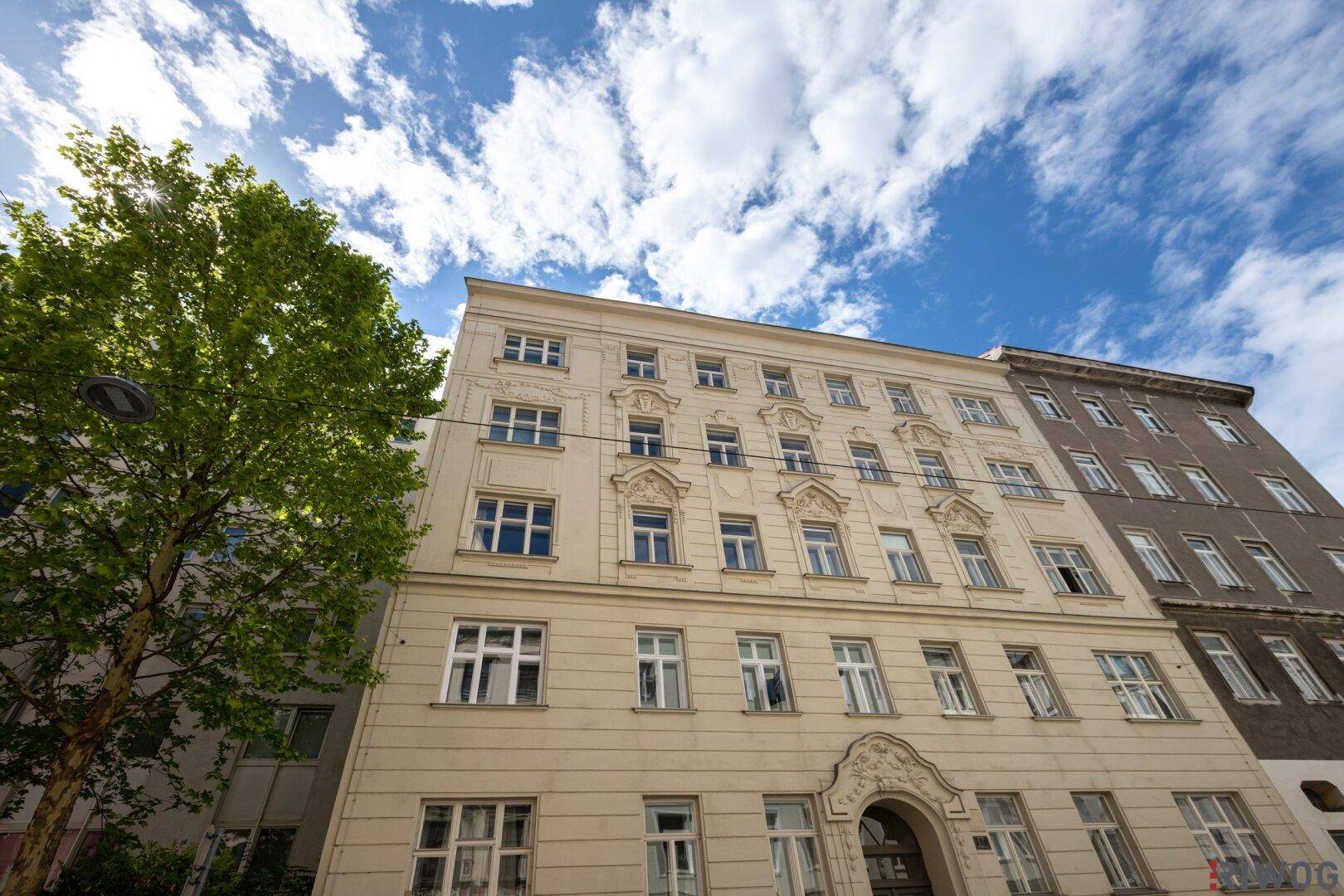 Klassische Fassade eines Altbaus unter blauem Himmel mit Baum im Vordergrund.