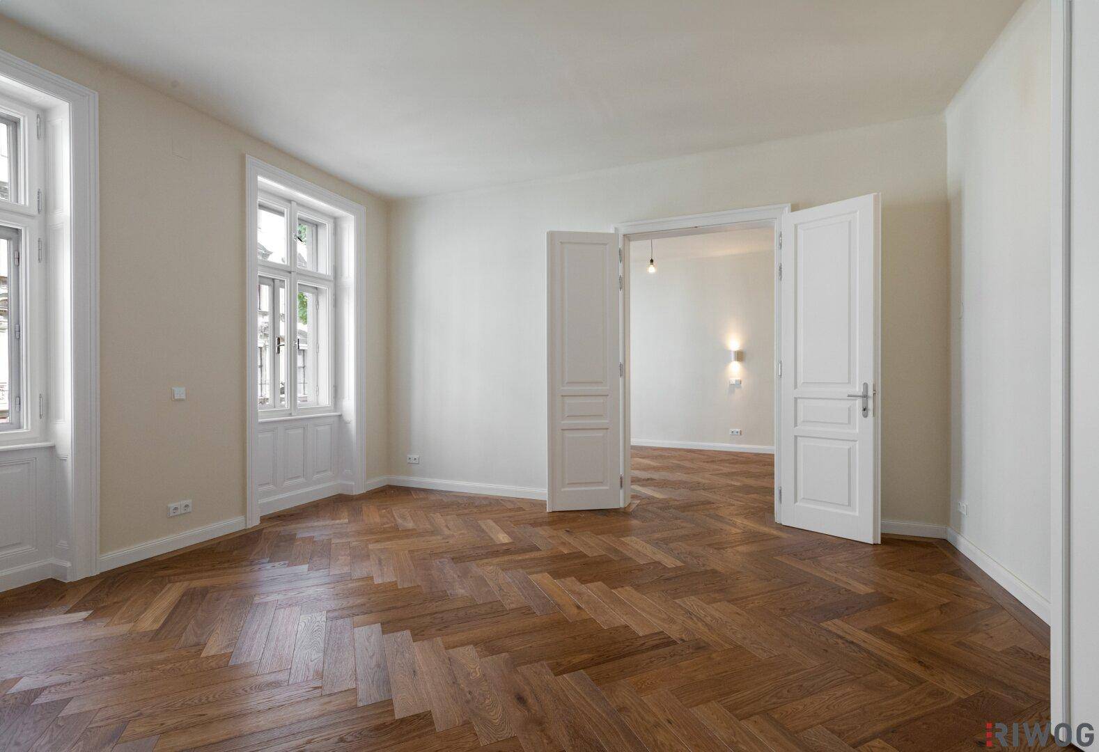 Geräumiges, leeres Wohnzimmer mit Fischgrätparkett und hohen Fenstern in einem Altbau.