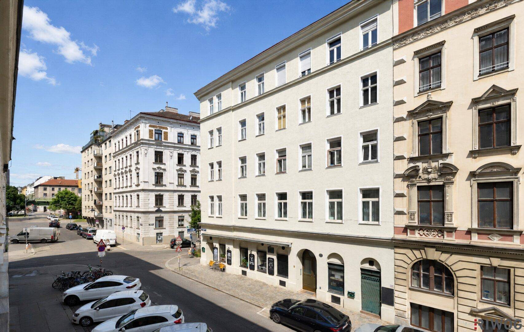 Ansicht einer belebten Stadtstraße mit mehreren historischen Wohngebäuden unter blauem Himmel.