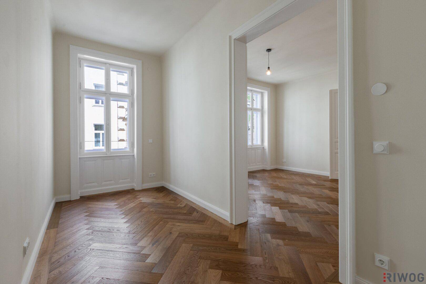 Helles Zimmer mit Fischgrätparkett und hohen Fenstern, das zu einem weiteren Raum führt.