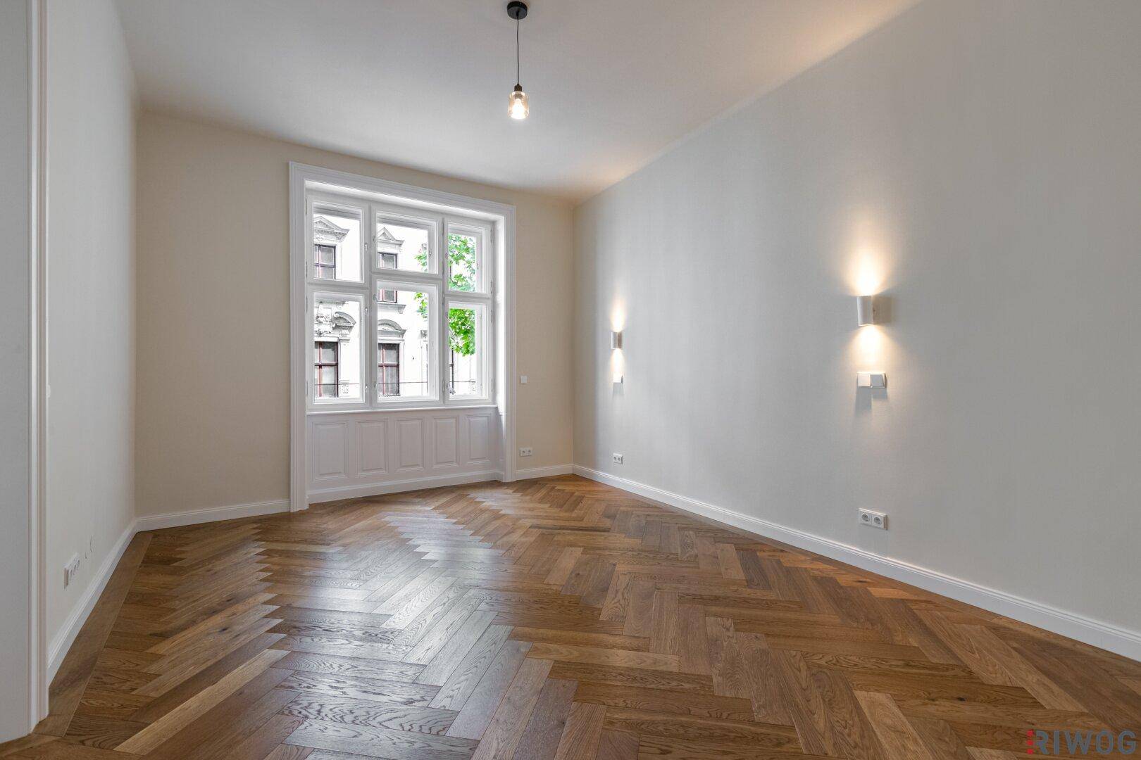 Helles, leeres Zimmer mit Fischgrätparkett, Wandleuchten und einem großen Fenster.