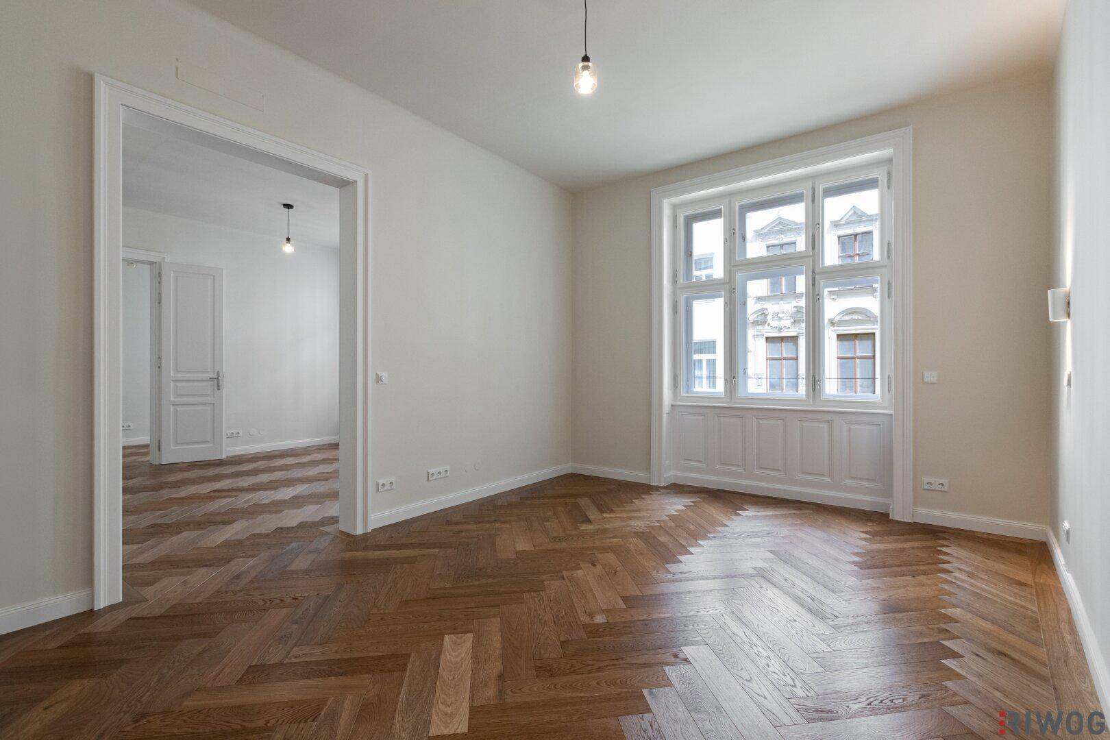 Geräumiges Zimmer mit Fischgrätparkett und hohen Fenstern, das zu einem weiteren Raum führt.