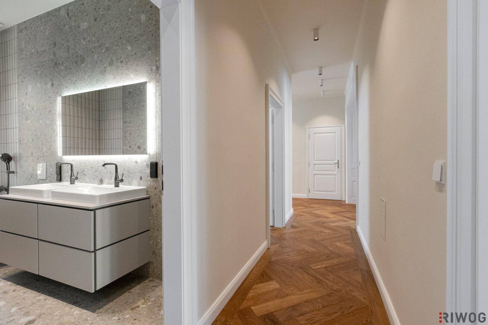 Flur mit Fischgrätparkett und Blick auf ein modernes Badezimmer mit Doppelwaschbecken.