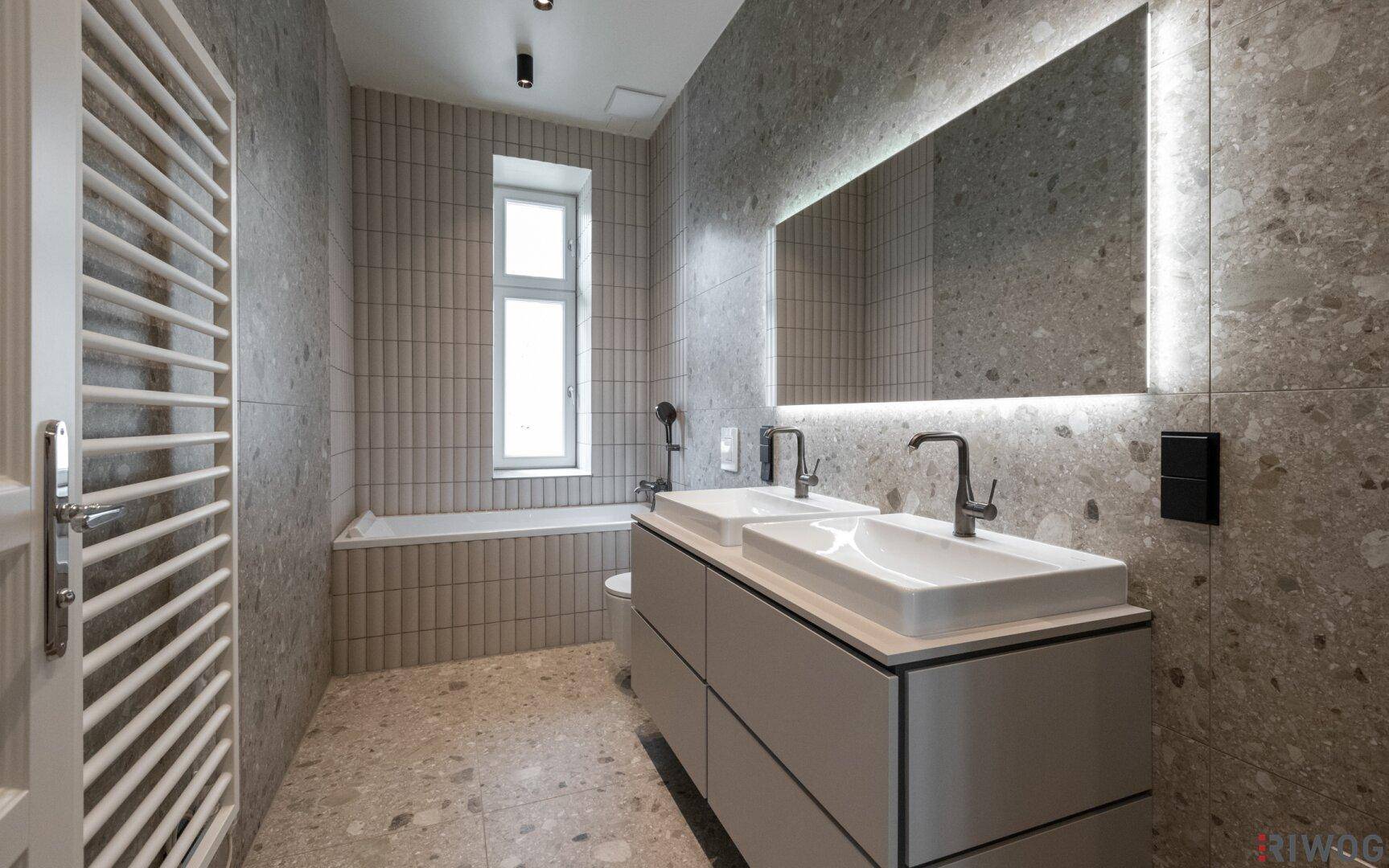 Modernes Badezimmer mit Doppelwaschbecken, beleuchtetem Spiegel und grauen Fliesen.