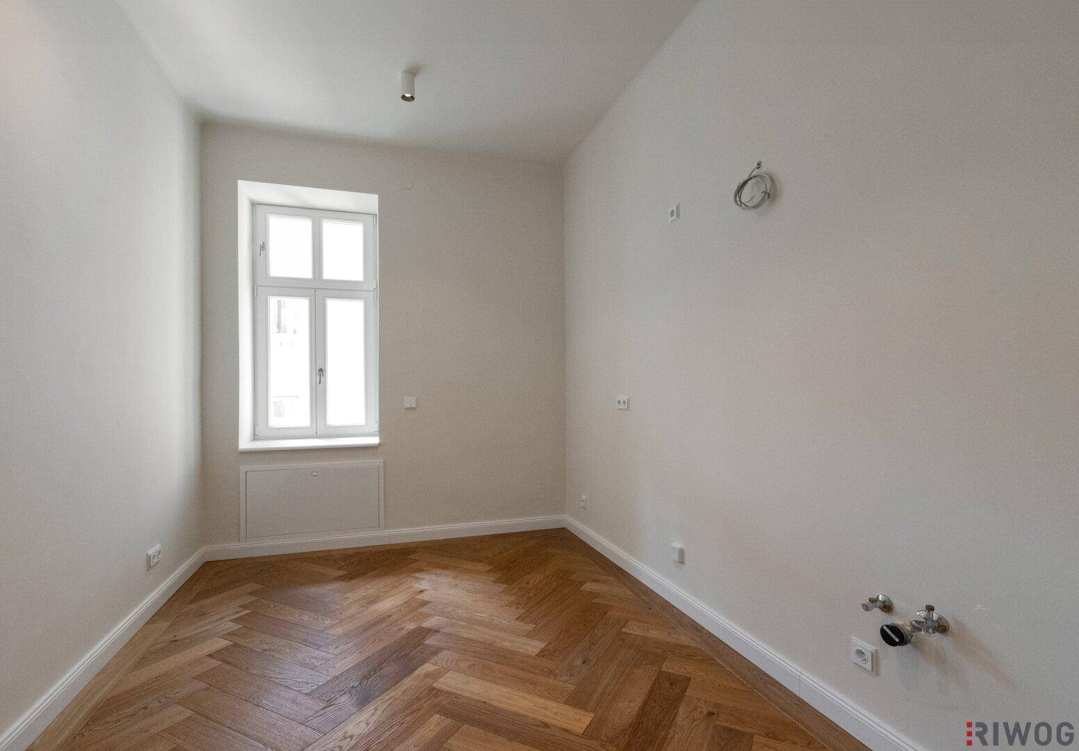Leerer Küchenraum mit Fischgrätparkett, Fenster und Anschlüssen für eine Einbauküche.