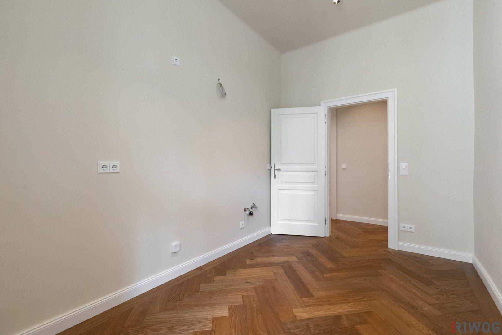 Leerer Küchenraum mit Fischgrätparkett, Fenster und Anschlüssen für eine Einbauküche.