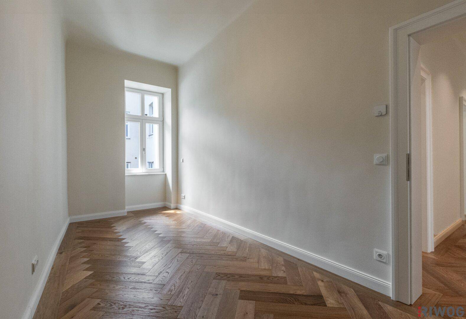 Helles, leeres Zimmer mit Fischgrätparkett und Fenster, ideal als Kinder- oder Arbeitszimmer.