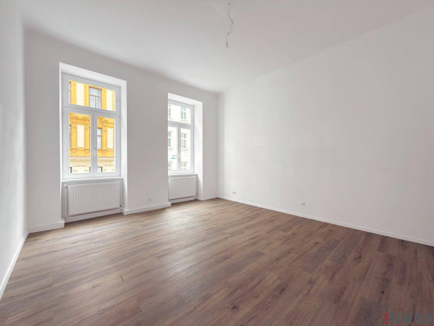 Geräumiger Raum mit Holzboden und zwei Fenstern, die eine helle Atmosphäre schaffen.