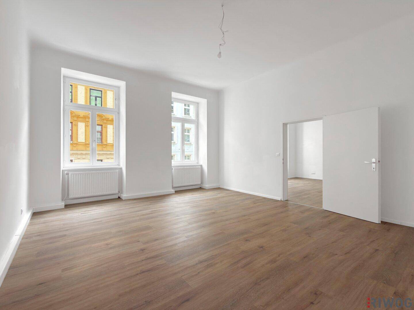 Heller Raum mit Holzboden und zwei Fenstern, die viel Tageslicht hereinlassen.
