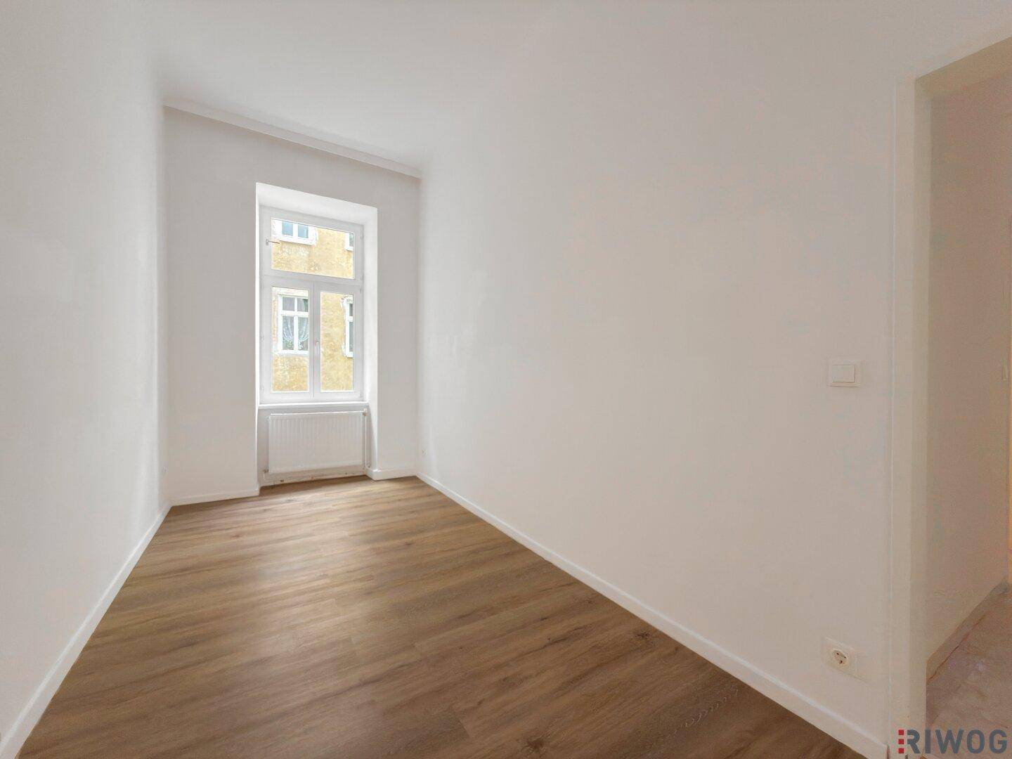 Kleinerer Raum mit Holzboden und einem Fenster, das Tageslicht hereinlässt.