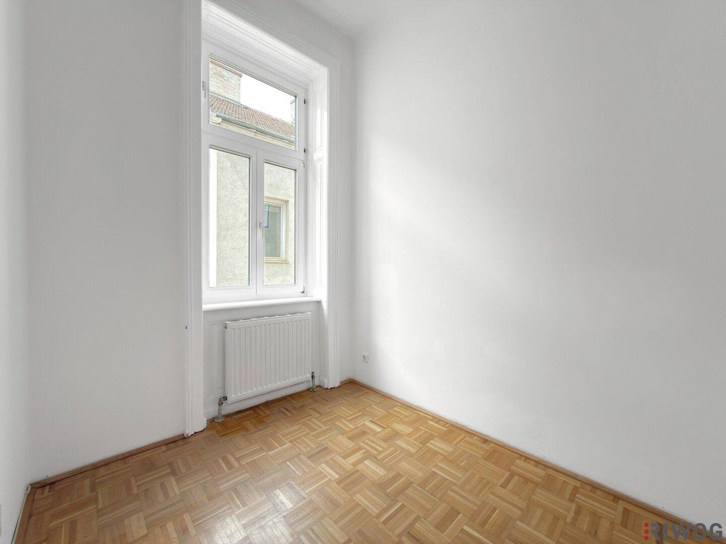 Heller, leerer Raum mit Parkettboden und einem Fenster, das viel Tageslicht spendet.