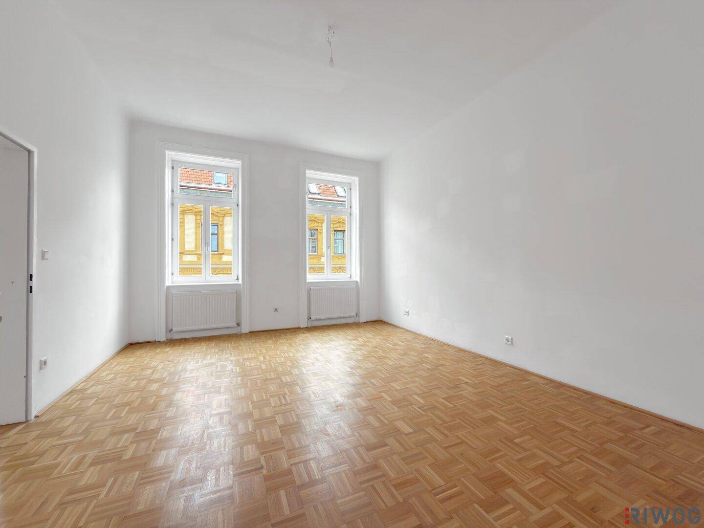 Geräumiger, leerer Raum mit Parkettboden und zwei Fenstern, die für gute Belichtung sorgen.