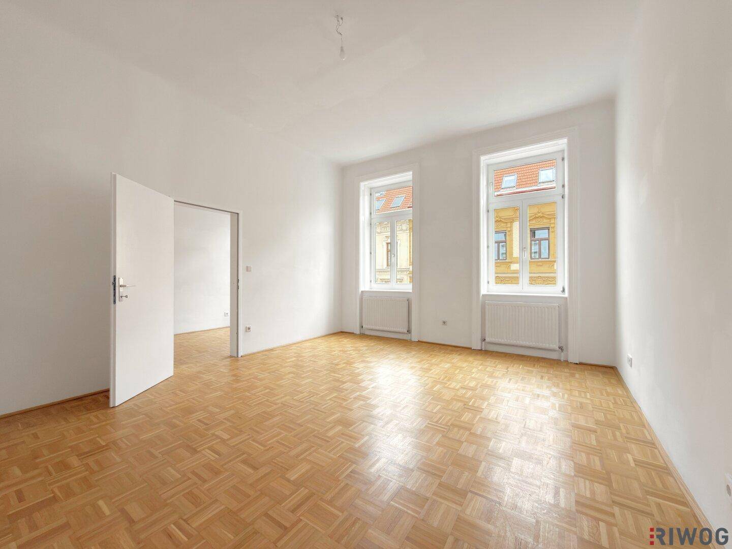 Leerer Raum mit Fischgrätparkettboden und zwei großen Fenstern, die viel Licht hereinlassen.