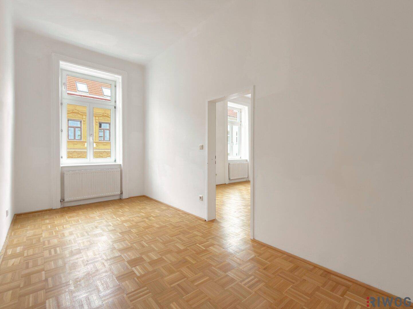 Heller, leerer Raum mit Parkettboden und einem Fenster, das in einen weiteren Raum führt.