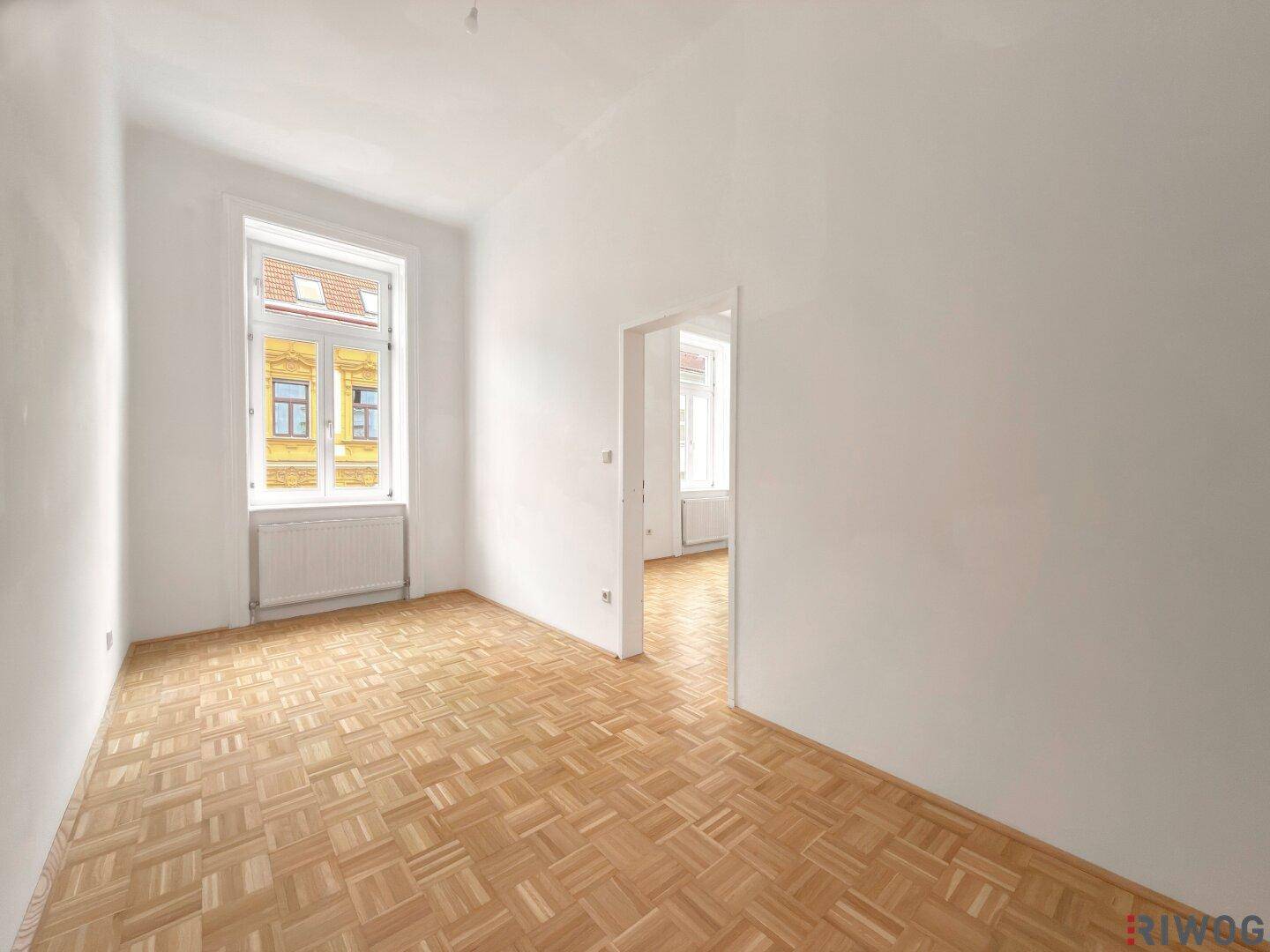 Heller, leerer Raum mit Parkettboden und einem Fenster, das in einen weiteren Raum führt.