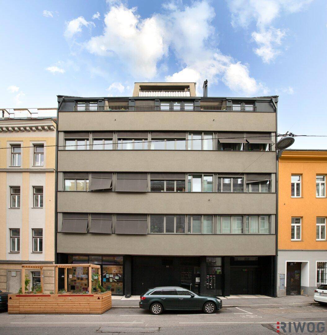 Moderne Gebäudefassade mit großen Fenstern und Jalousien an einer städtischen Straße, mit geparkten Autos davor.