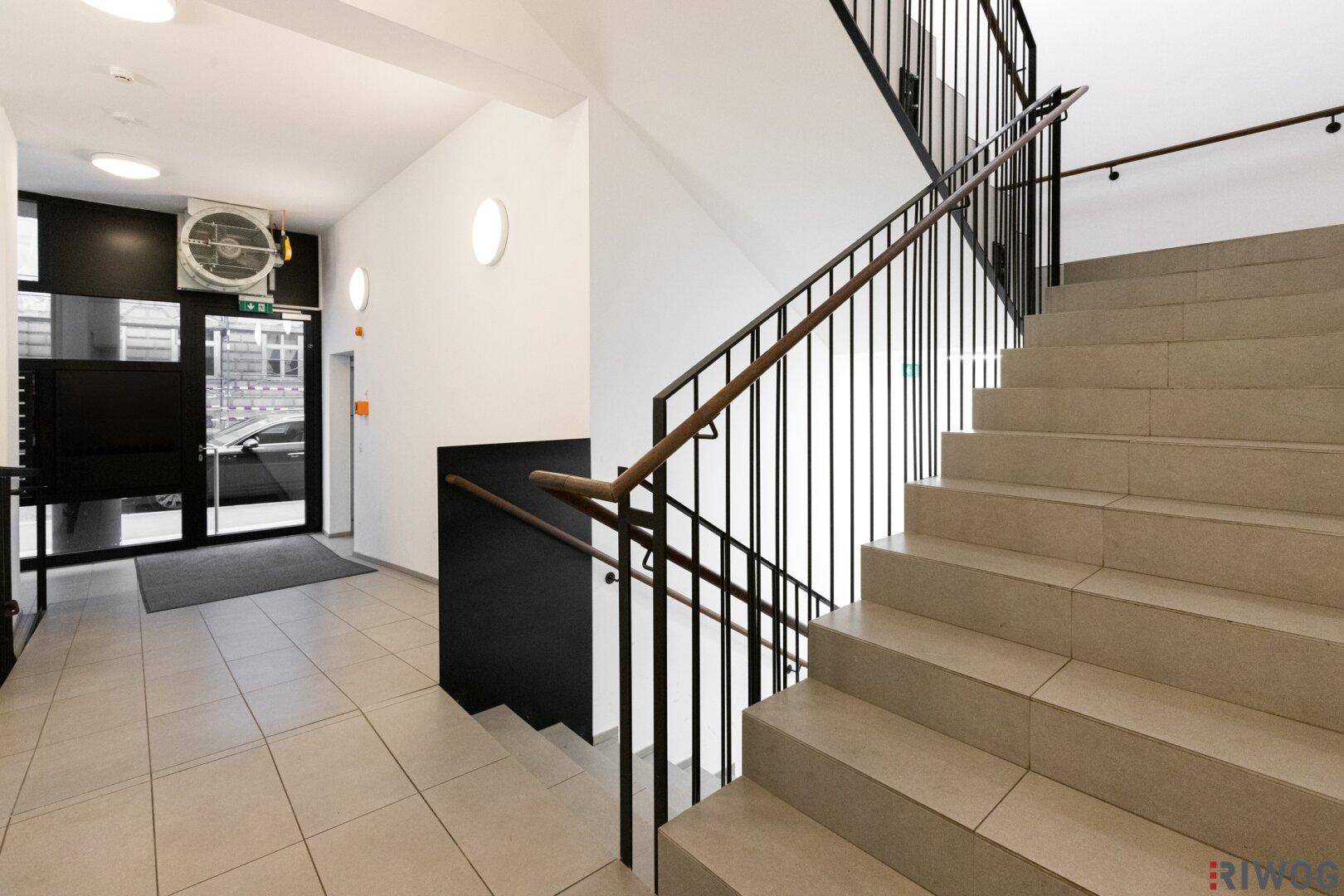 Heller Eingangsbereich mit gefliestem Boden und einer Treppe mit schwarzem Geländer und Holzhandlauf.