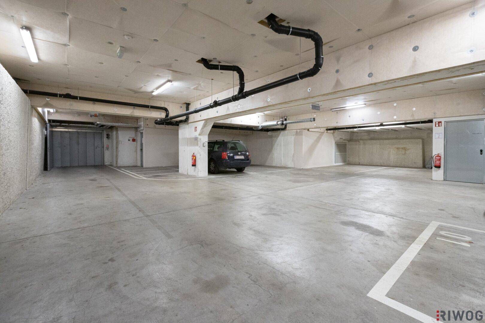 Weitläufige Tiefgarage mit vielen Stellplätzen, Betonwänden und Deckeninstallationen, gut beleuchtet.