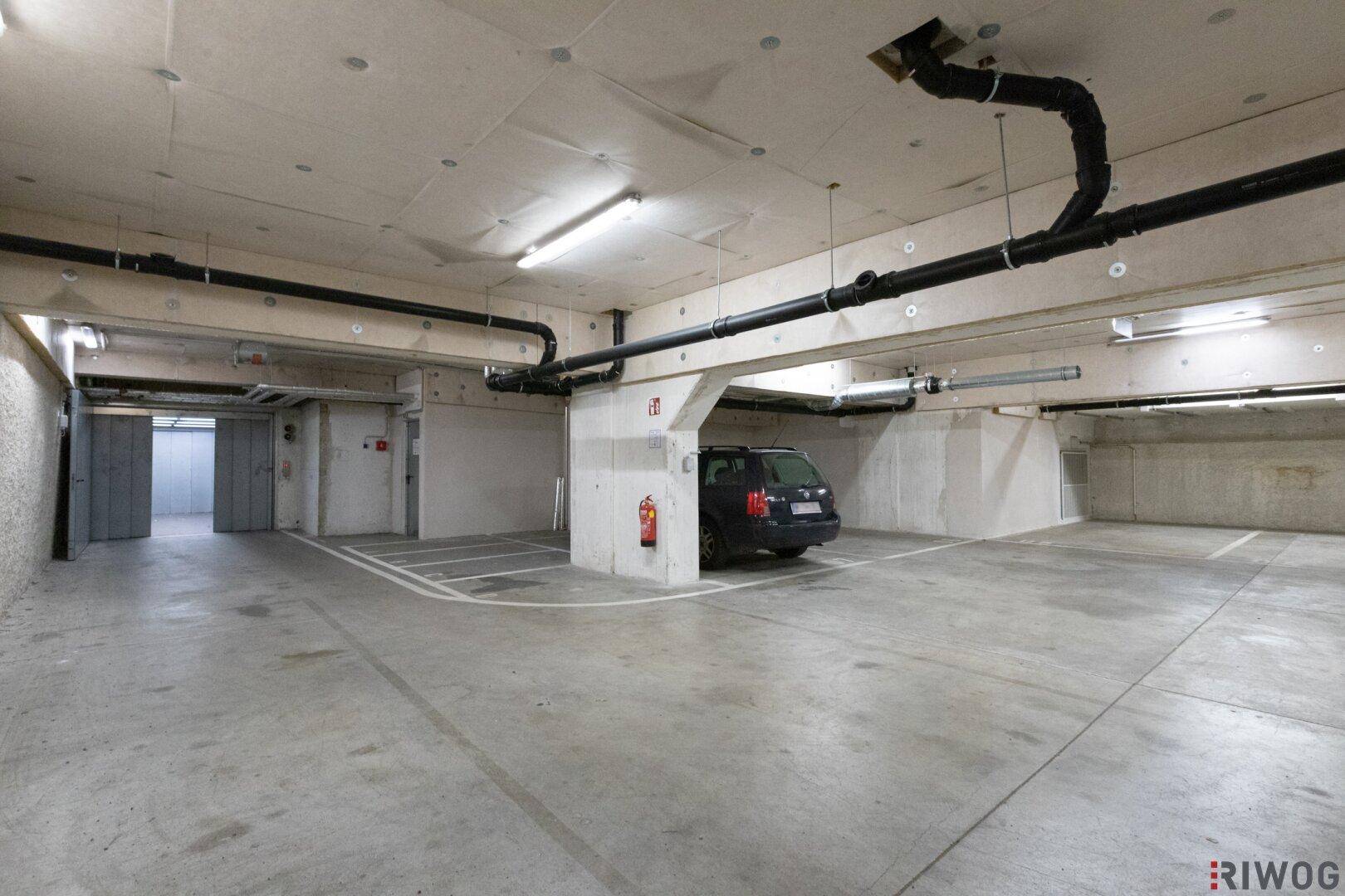 Ansicht der Tiefgarage mit Betonstützen, Stellplätzen und sichtbaren Versorgungsleitungen an der Decke.